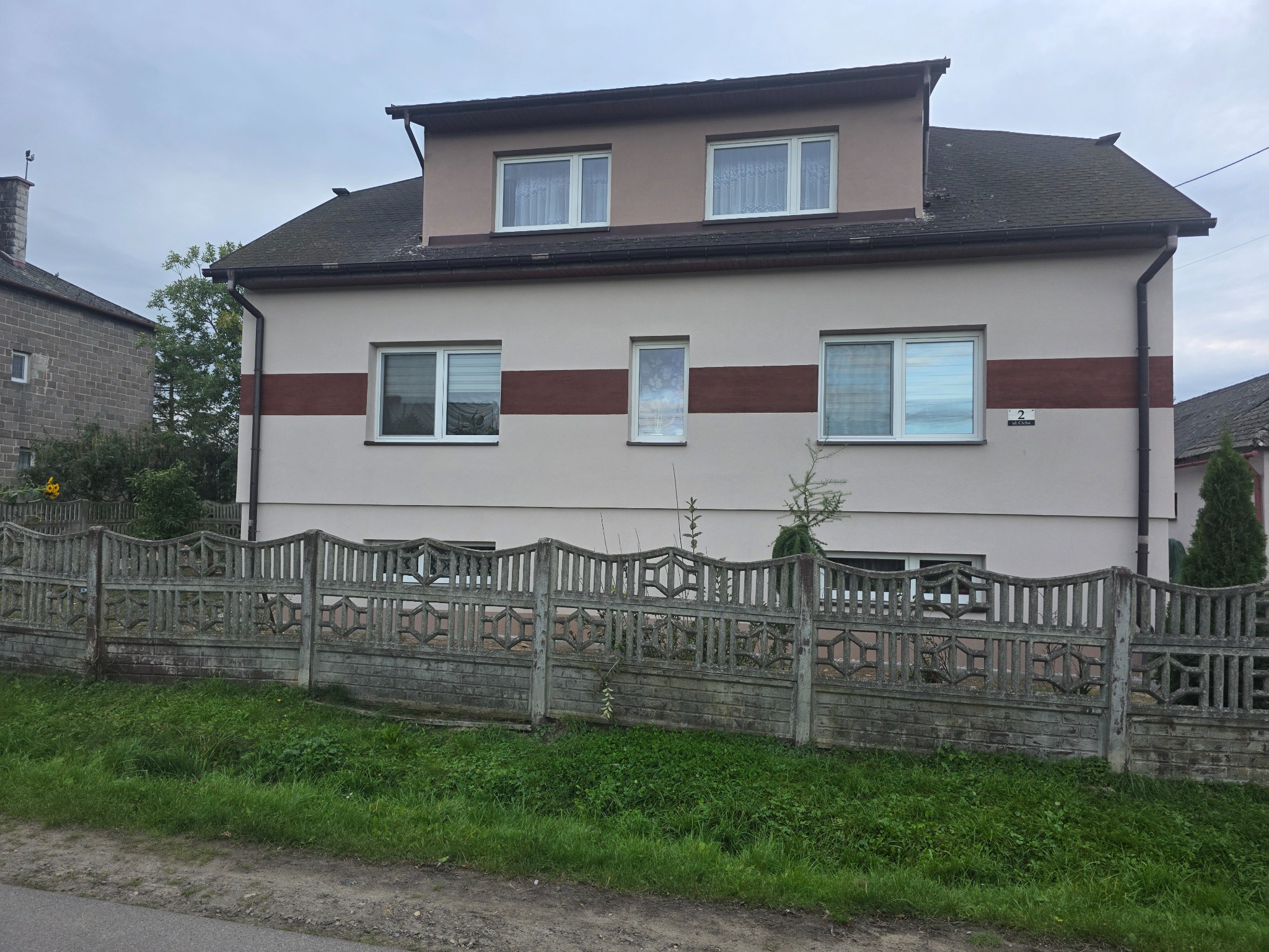 Dwukondygnacyjny dom z beżową elewacją i brązowym pasem. Widoczny betonowy płot i trawnik. Pochmurne niebo w tle.