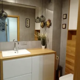 Nowoczesna łazienka z szarymi płytkami, drewnianą podłogą i blatem, białą szafką pod umywalką oraz wiszącymi lampami z szarego szkła, dekorowana ceramicznymi rybami i ramkami ze zdjęciami na ścianie.