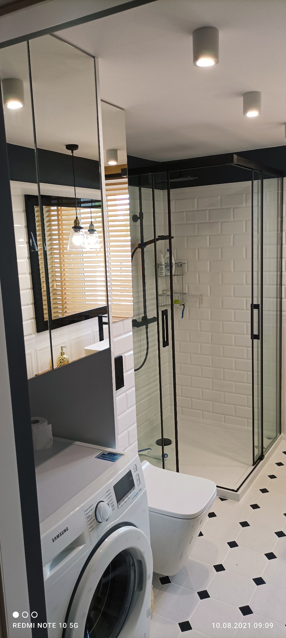 Nowoczesna łazienka z kabiną prysznicową w stylu loft, białe płytki typu metro, czarne ramy kabiny, pralka Samsung i lustro z odbiciem żaluzji okiennych.