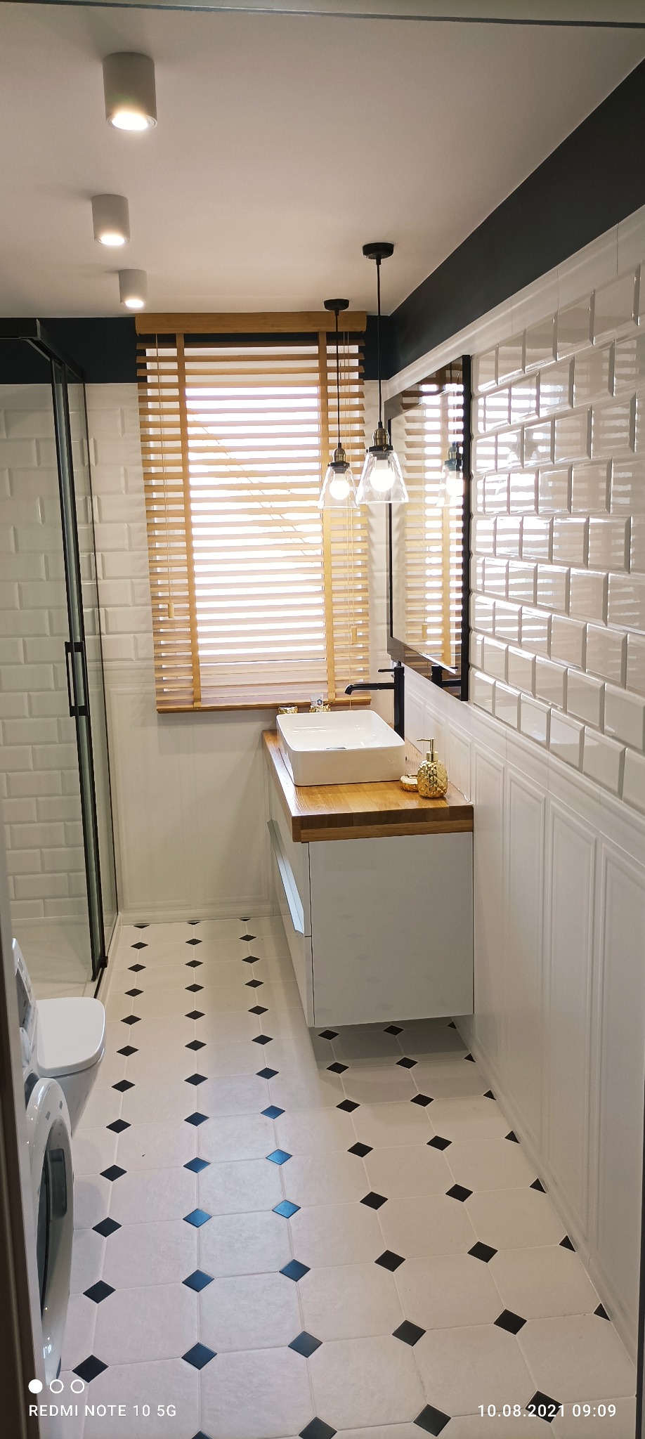 Nowoczesna łazienka w bieli i czerni z drewnianymi akcentami: białe płytki imitujące cegłę, biała umywalka nablatowa na drewnianym blacie, lustro w czarnej ramie, drewniane żaluzje i podłoga...