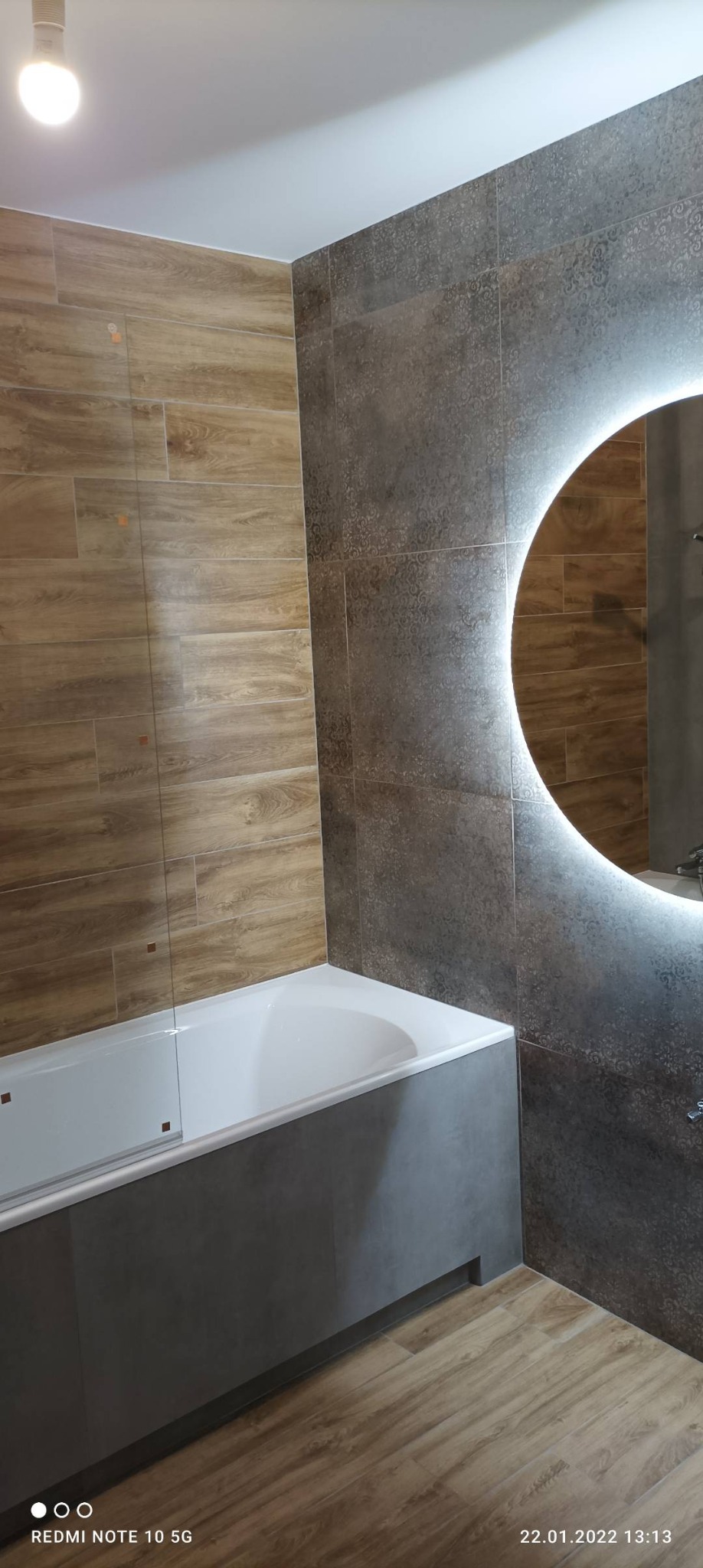 Nowoczesna łazienka z drewnopodobnymi płytkami na ścianach i podłodze, szarą obudową wanny i okrągłym lustrem z podświetleniem LED.