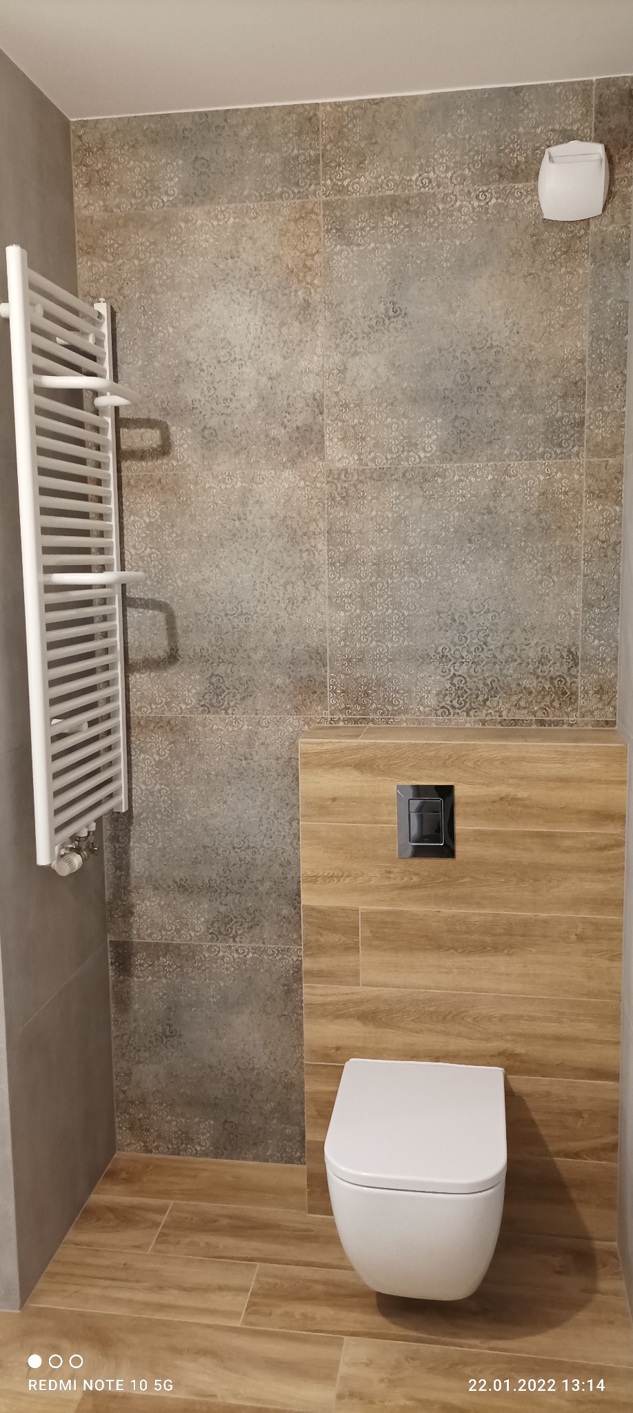 Nowoczesna łazienka z wiszącą toaletą, grzejnikiem drabinkowym i płytkami imitującymi drewno na podłodze i części ściany, ozdobne płytki na pozostałej części ściany.