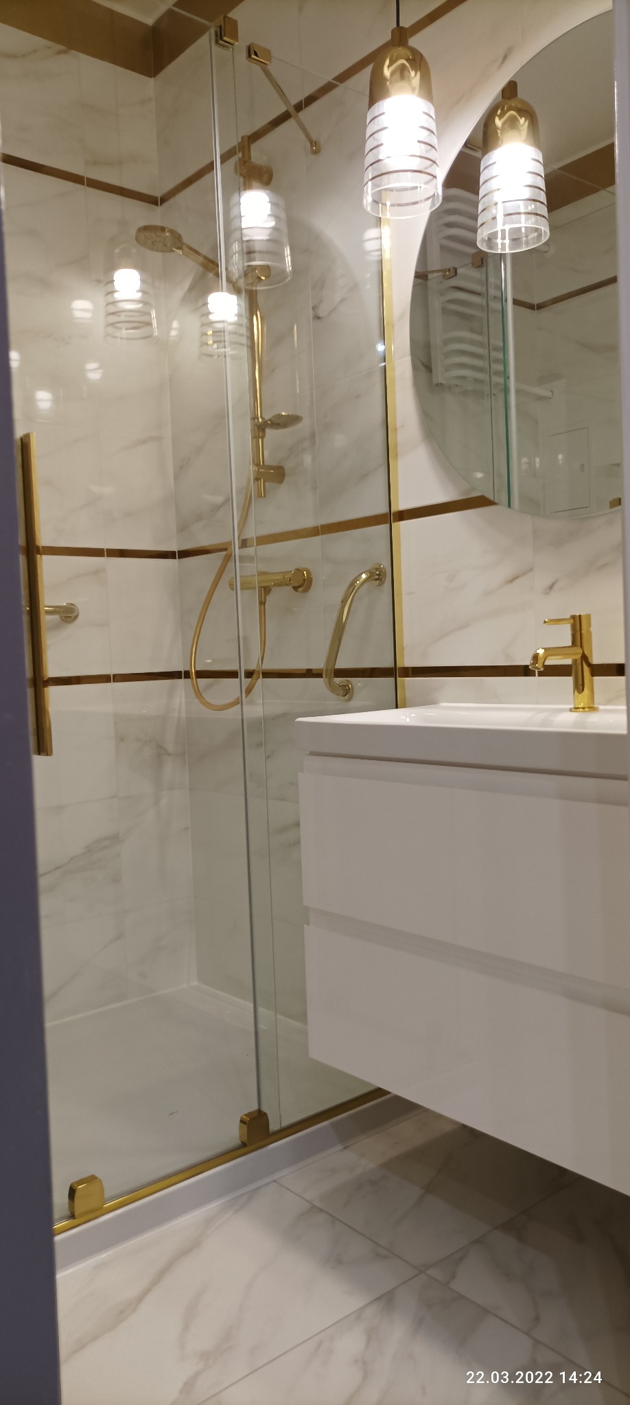 Nowoczesna łazienka z kabiną prysznicową ze złotymi detalami, białą umywalką z szafką i marmurowymi płytkami na ścianach i podłodze, oświetlona wiszącymi lampami.