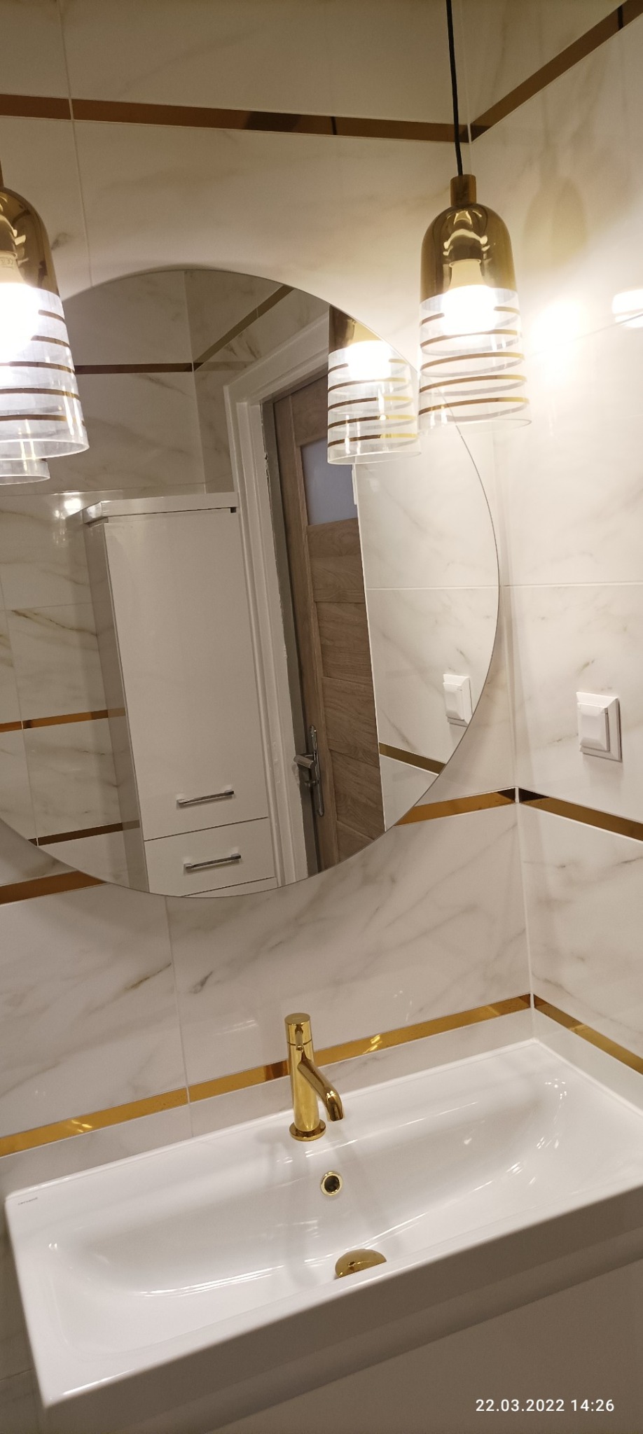 Nowoczesna łazienka z marmurowymi płytkami, złotymi akcentami i okrągłym lustrem odbijającym szafkę i drzwi.