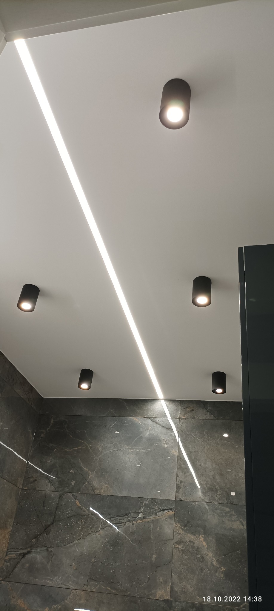 Nowoczesna łazienka z ciemnymi płytkami imitującymi marmur, podświetlonym sufitem z listwą LED i czarnymi, punktowymi oprawami oświetleniowymi.