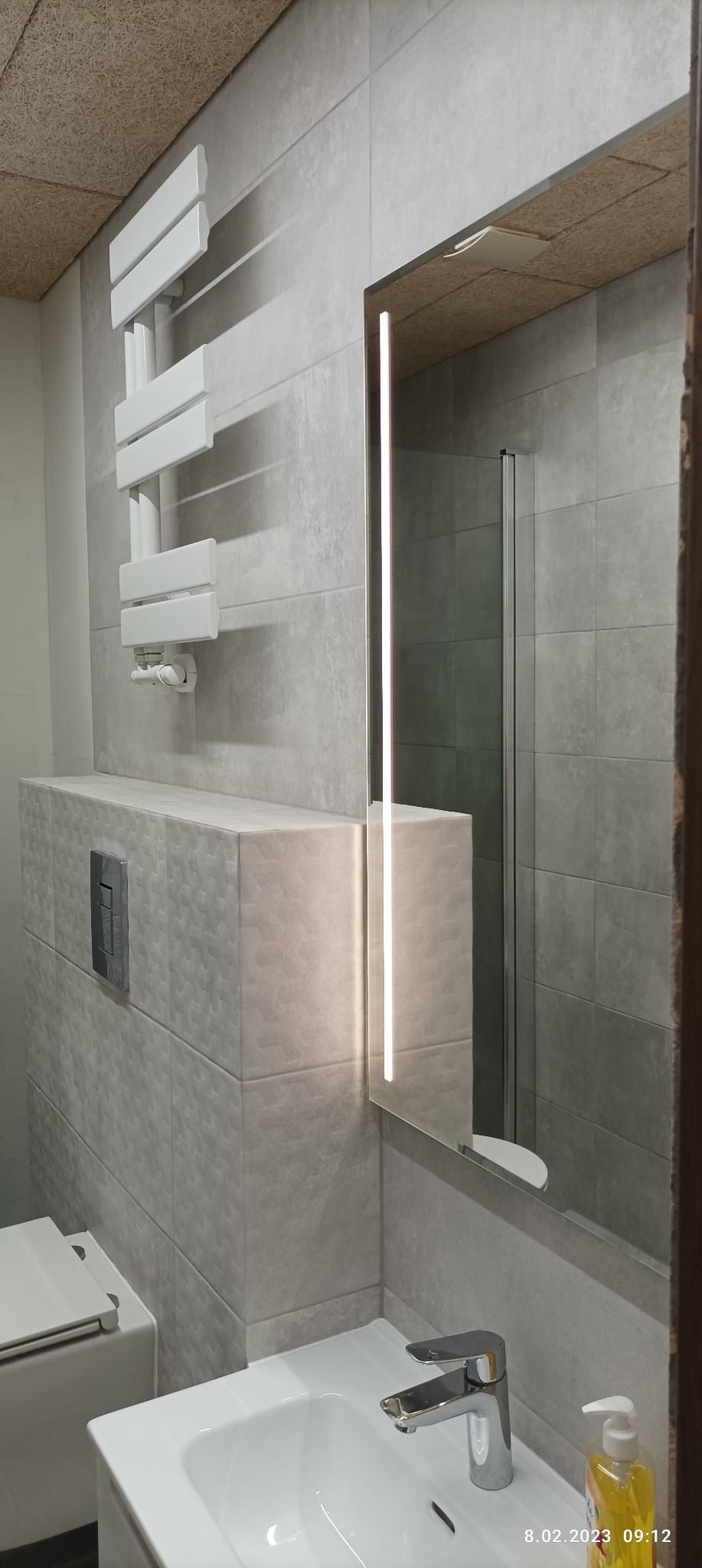 Szara łazienka z podświetlanym lustrem, grzejnikiem drabinkowym i umywalką z baterią, widoczna kabina prysznicowa w tle, sufit wykończony panelami z naturalnego drewna.