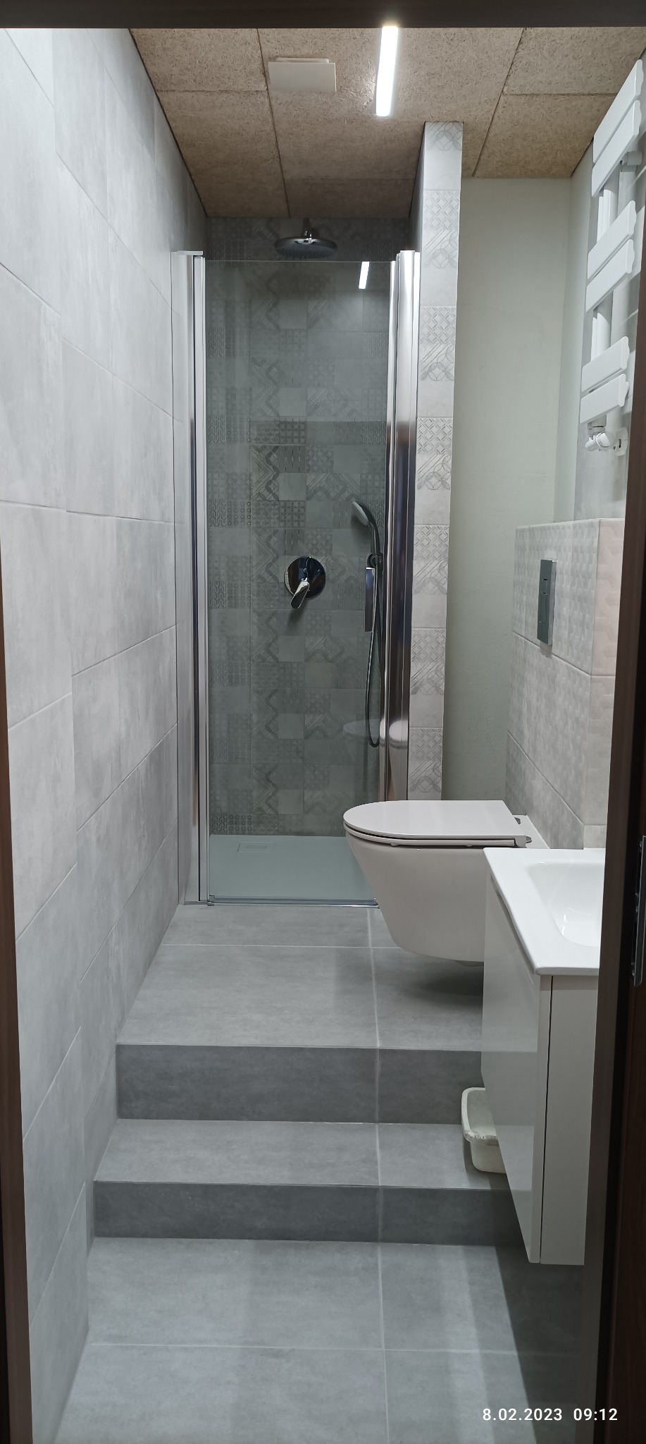 Wykończona łazienka z kabiną prysznicową z wzorzystą ścianą, podestem z płytek imitujących schody, podwieszaną toaletą i białą szafką pod umywalką, oświetleniem LED na suficie z płyty OSB.