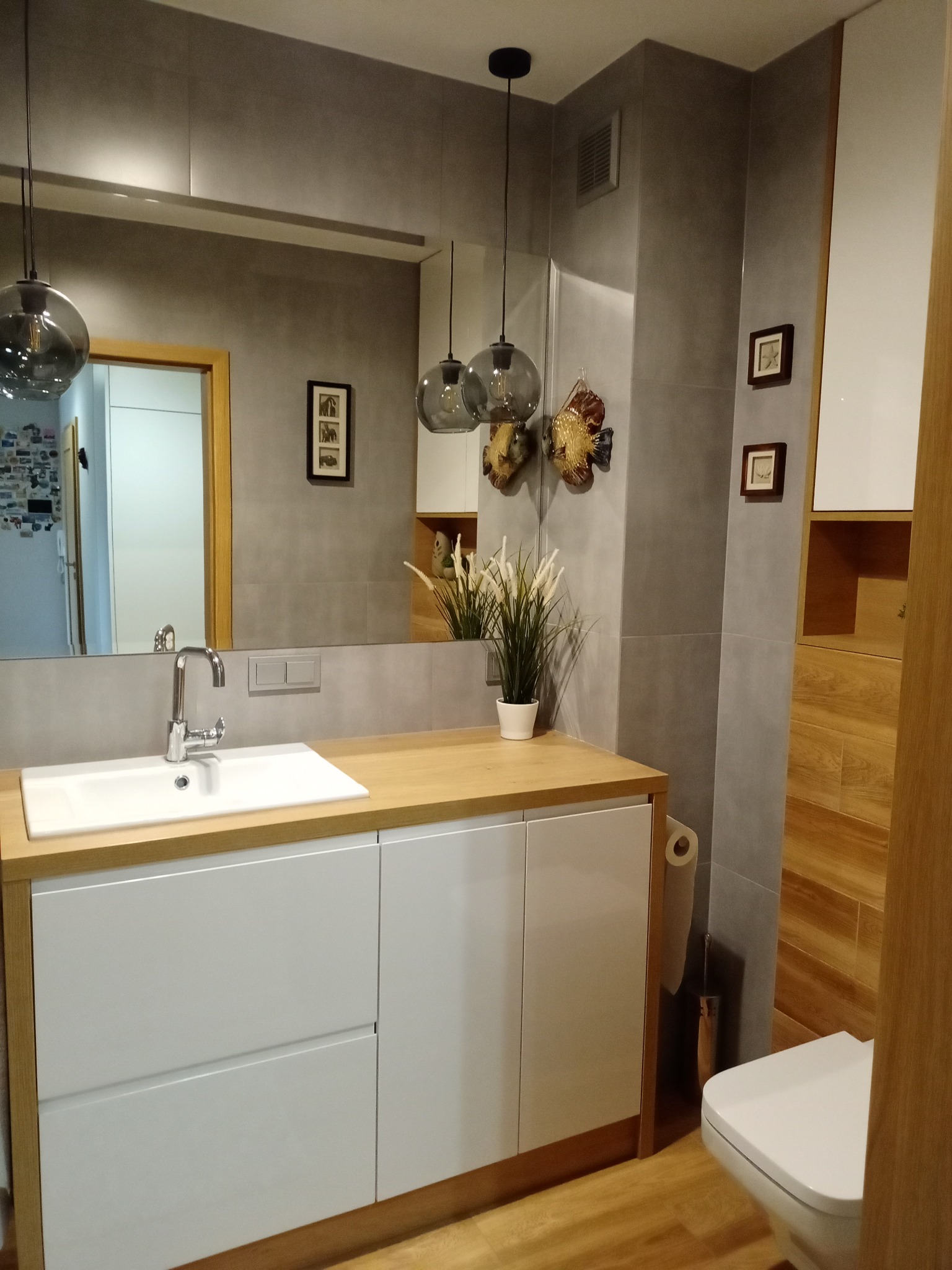 Nowoczesna łazienka z szarymi płytkami, drewnianą podłogą i blatem, białą szafką pod umywalką oraz wiszącymi lampami z szarego szkła, dekorowana ceramicznymi rybami i ramkami ze zdjęciami na ścianie.