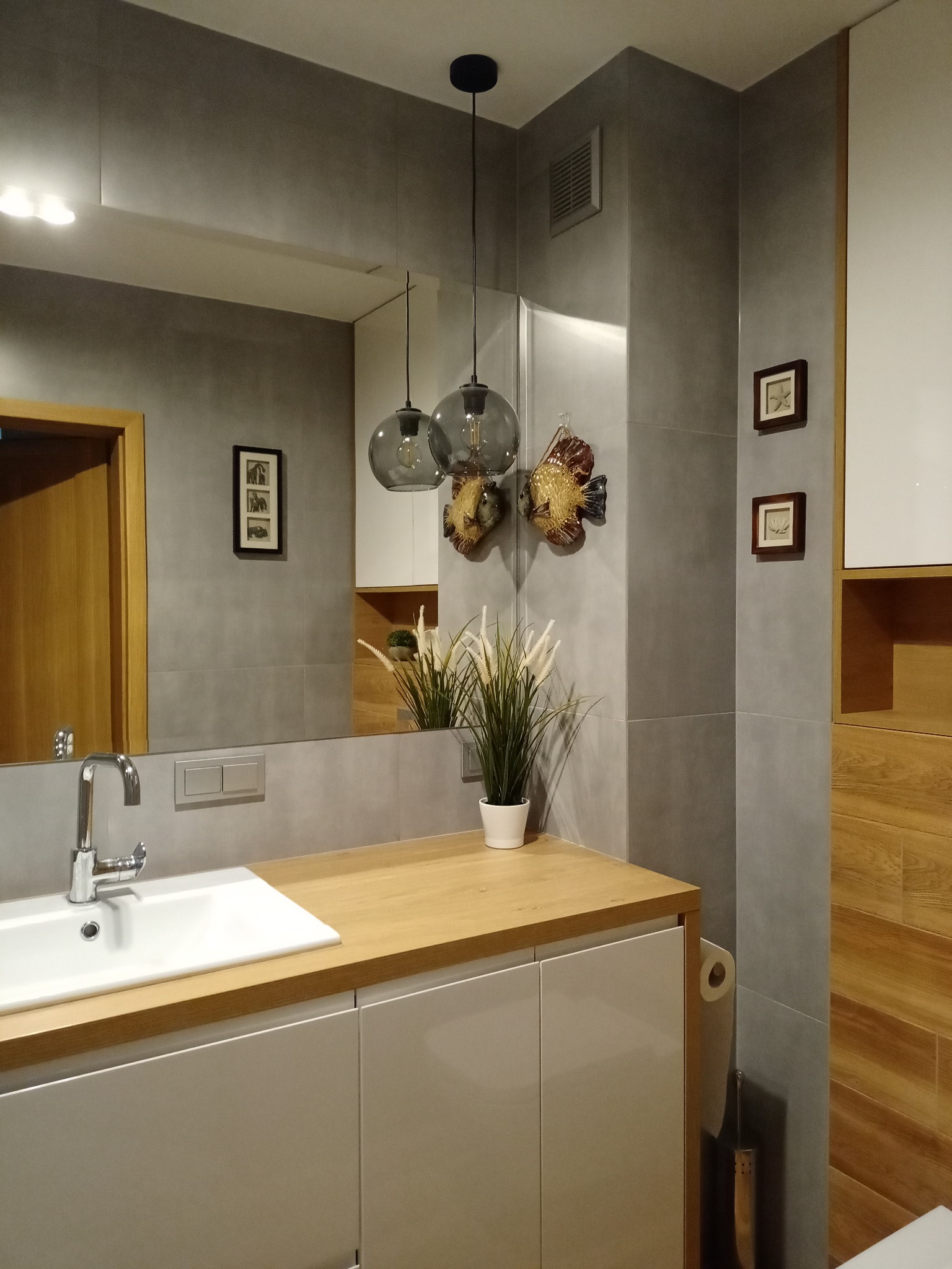Nowoczesna łazienka z szarą glazurą, drewnianym blatem, białymi szafkami pod umywalką i dekoracyjnymi, szklanymi lampami wiszącymi w kształcie kul. Na ścianie wiszą ozdobne ceramiczne ryby i małe...