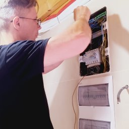 Kranikontakt - Mężczyzna w okularach podłącza kolorowe przewody w otwartej rozdzielnicy elektrycznej, widoczne drewniane belki sufitu i czerwone peszle.