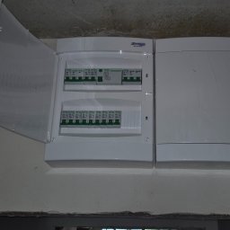 Kranikontakt - Nowa rozdzielnica elektryczna z otwartą obudową, ukazująca zielone wyłączniki GACIA i SCHRACK, zamontowana na surowej ścianie w trakcie budowy. Obok zamknięta skrzynka.