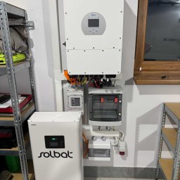 MCE MM - Zamontowany biały inwerter Deye i magazyn energii Solbat na ścianie w garażu. Widoczne kable i skrzynki elektryczne. Obok regał metalowy.