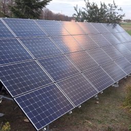 MCE MM - Panel słoneczny z widocznym odbiciem nieba, zamontowany na metalowej konstrukcji, na tle zieleni i krajobrazu wiejskiego.