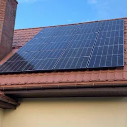 Panel słoneczny na dachu z czerwoną dachówką, obok ceglany komin. Błękitne niebo w tle. Widok z dołu na dom z zamontowaną instalacją fotowoltaiczną.