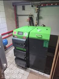 Zielono-czarny piec na paliwo stałe MCE w kotłowni z szarymi płytkami i instalacją rurową. Widoczne zasobniki na paliwo i panel sterowania pieca.