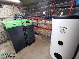 Kocioł na paliwo stałe MCE z zielonym akcentem obok białego zasobnika ciepłej wody w kotłowni z surowymi, szarymi ścianami z betonu komórkowego.