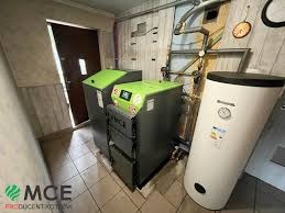 Nowoczesny piec i zasobnik w kotłowni w Radzyniu Podlaskim. Zielone akcenty na obudowie pieca, widoczne rury i zegar na ścianie. Kompaktowe rozwiązanie grzewcze.