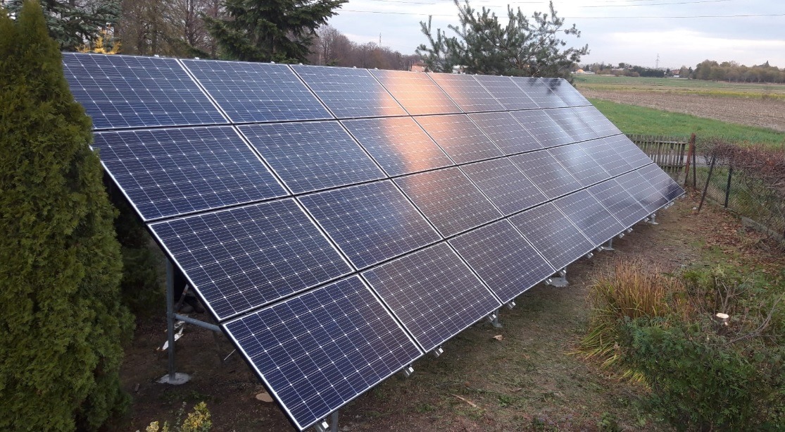 Panel słoneczny z widocznym odbiciem nieba, zamontowany na metalowej konstrukcji, na tle zieleni i krajobrazu wiejskiego.