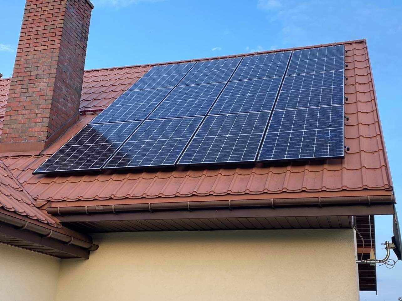 Panel słoneczny na dachu z czerwoną dachówką, obok ceglany komin. Błękitne niebo w tle. Widok z dołu na dom z zamontowaną instalacją fotowoltaiczną.