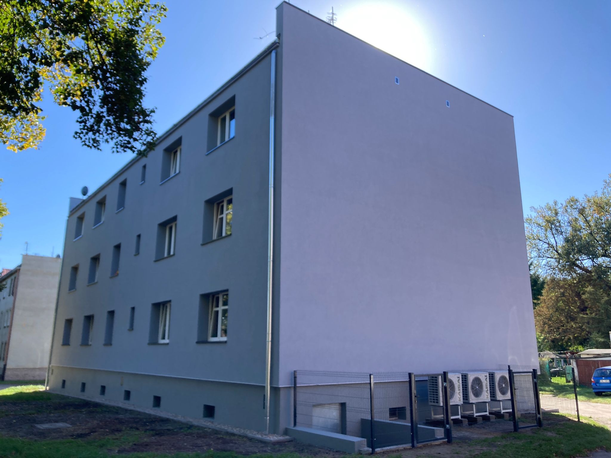 Odnowiona elewacja bloku mieszkalnego w odcieniach szarości z zainstalowanymi klimatyzatorami, oświetlona jasnym słońcem i błękitnym niebem. Widok z dołu.