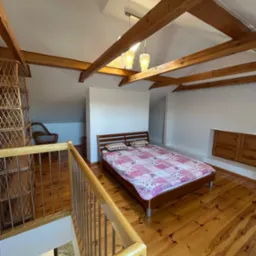 Pokój na poddaszu z drewnianą podłogą, odsłoniętymi belkami stropowymi, łóżkiem z patchworkową narzutą i wiklinowym regałem, widziany z góry z poziomu balustrady schodów.