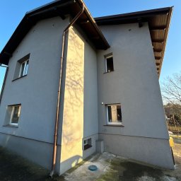 Goldbud - Dwukondygnacyjny dom z szarą elewacją, brązowymi rynnami i drewnianą podbitką dachową, widziany pod kątem w słoneczny dzień.