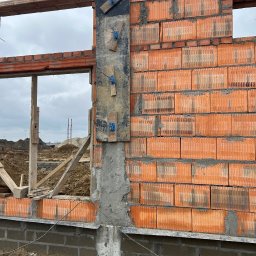 Goldbud - Wznoszenie ściany z czerwonej cegły ceramicznej z widocznymi otworami okiennymi, z użyciem drewnianych elementów szalunkowych i metalowych kotew, na tle placu budowy i pochmurnego nieba.