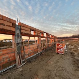 Goldbud - Wznoszenie muru z czerwonej ceramiki poryzowanej na fundamencie w trakcie budowy domu, widoczne rusztowania i paleta z bloczkami, tło stanowi zachodzące słońce i częściowo zachmurzone niebo.