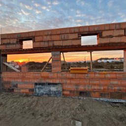 Goldbud - Wznoszone ściany domu murowanego z czerwonej cegły ceramicznej w trakcie budowy, widoczne otwory okienne i słupki konstrukcyjne, na tle zachodzącego słońca i częściowo zachmurzonego nieba.
