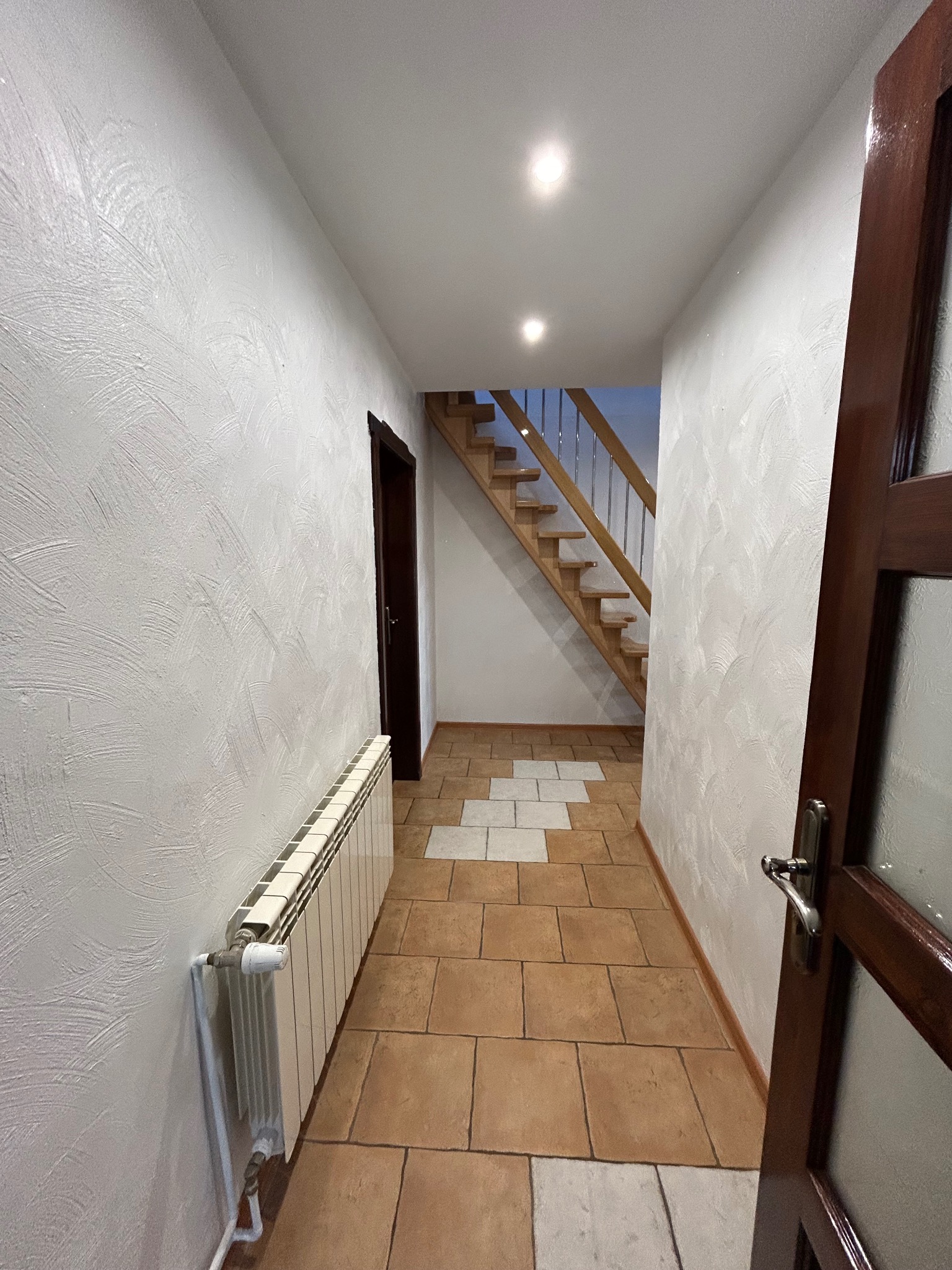 Wnętrze korytarza z drewnianymi schodami, brązową podłogą z płytek i grzejnikiem przy teksturowanej, białej ścianie. Widoczne drzwi z ciemnego drewna i przeszklonym panelem.