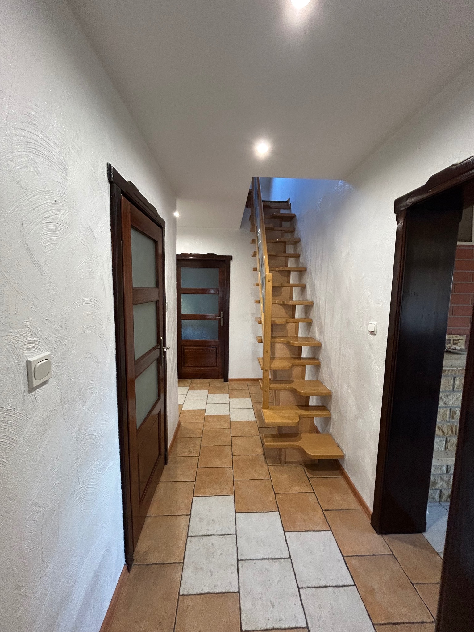 Wnętrze domu z drewnianymi schodami bez podstopnic, brązowymi drzwiami z przeszkleniami i podłogą w brązowo-białą szachownicę.