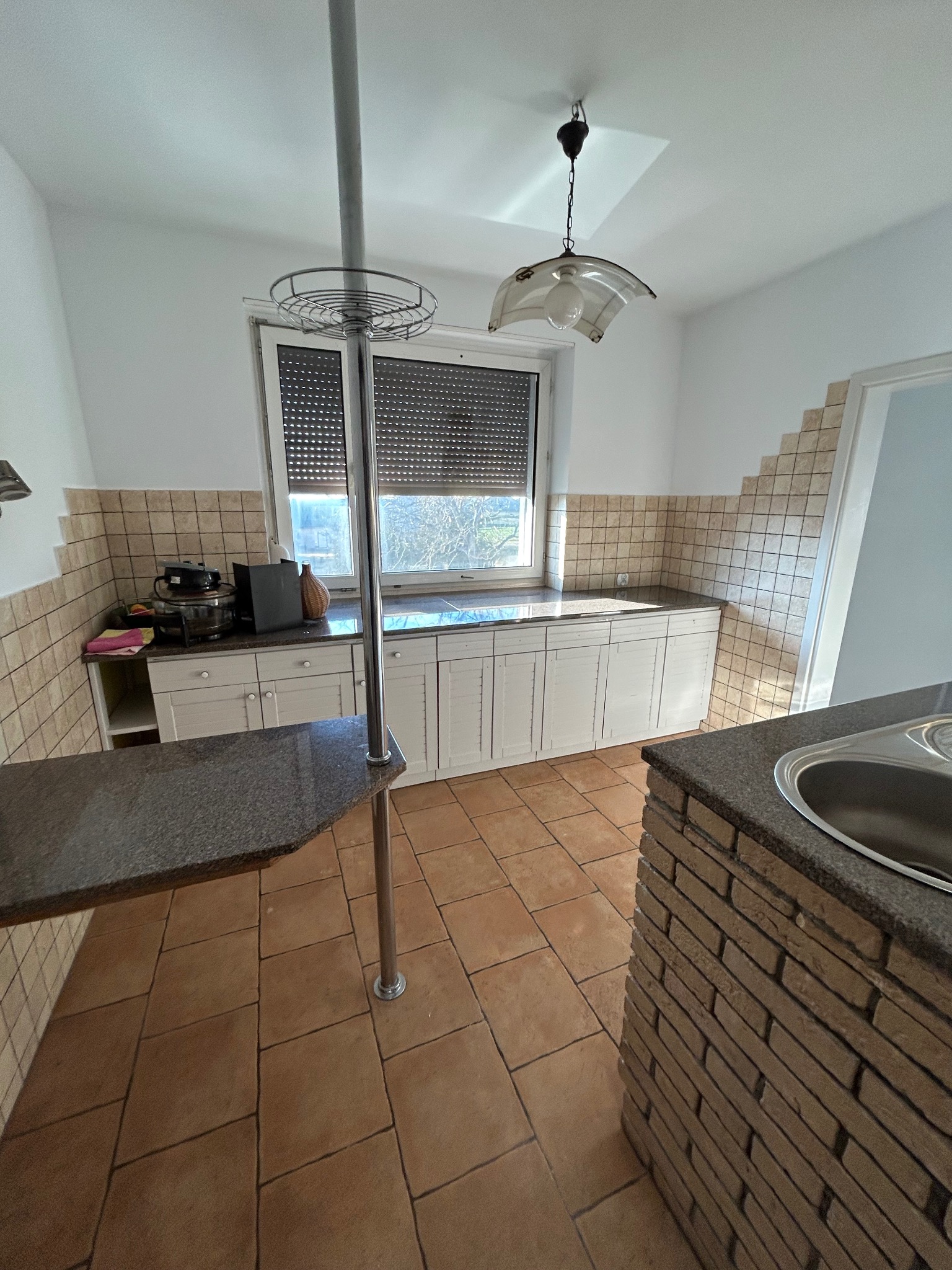 Wnętrze kuchni z brązową terakotą na podłodze, białymi szafkami, szarą granitową wyspą z chromowanym słupkiem i okapem wykończonym płytkami na ścianie.