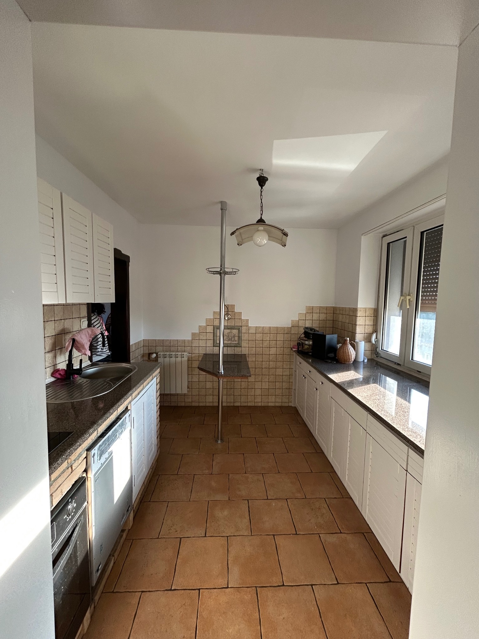 Jasna kuchnia z terakotową podłogą, białymi szafkami i granitowymi blatami, widok na okno i metalowy słupek z półkami.