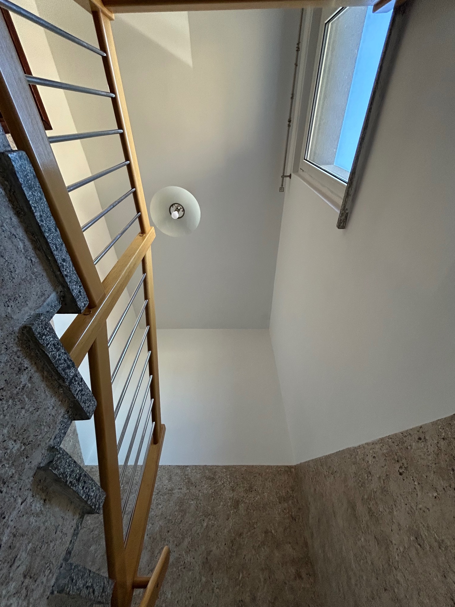 Wnętrze klatki schodowej z drewnianą balustradą ze stalowymi prętami, widok z dołu na sufit z lampą i okno dachowe w jasnym pomieszczeniu.