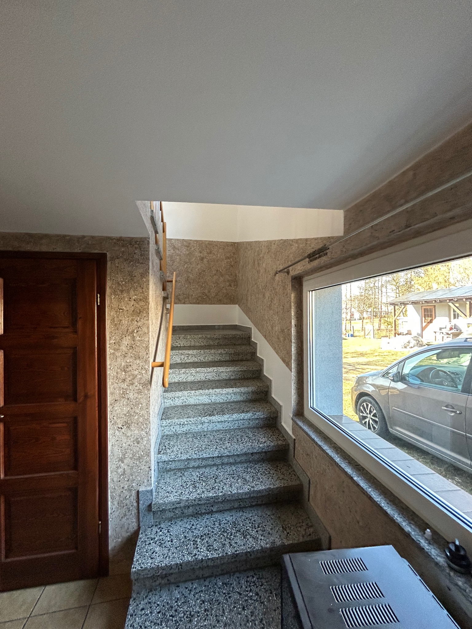 Wnętrze domu: granitowe schody z drewnianą balustradą, brązowe drzwi po lewej stronie, okno z widokiem na podwórko i szary samochód na zewnątrz.