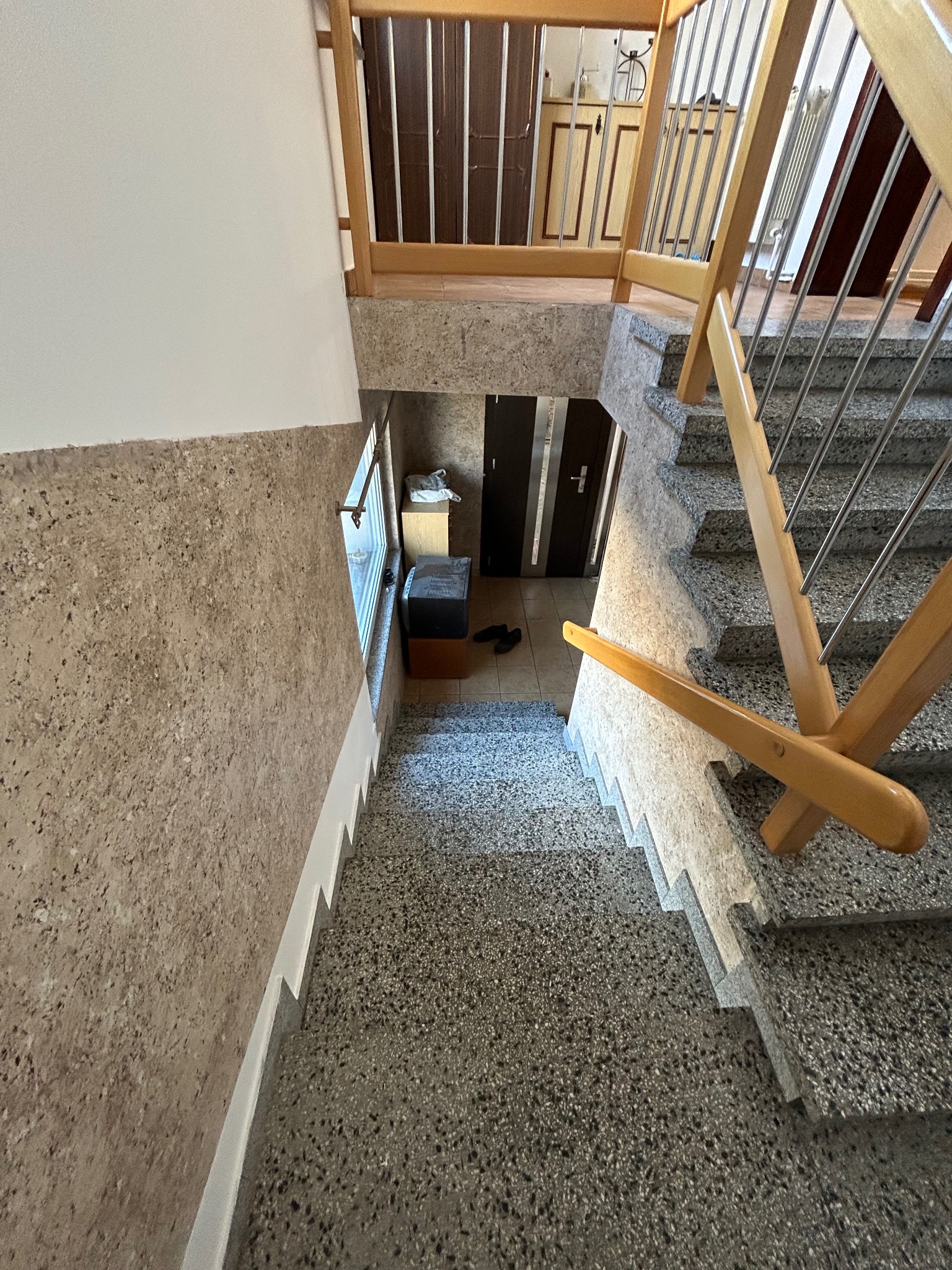 Widok z góry na klatkę schodową z lastryko, jasne drewniane poręcze ze stalowymi elementami, ściana z fakturą, okno z widocznym wyposażeniem korytarza i drzwiami.