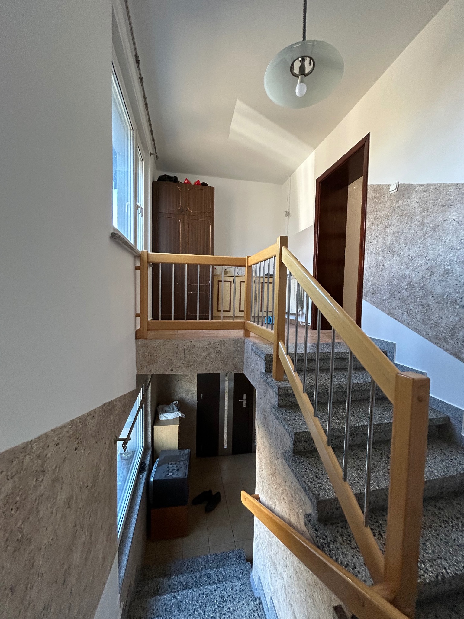 Wnętrze klatki schodowej w domu, z drewnianą balustradą z metalowymi prętami, schody wyłożone lastryko, widoczna szafa w zabudowie na półpiętrze i wisząca lampa sufitowa.