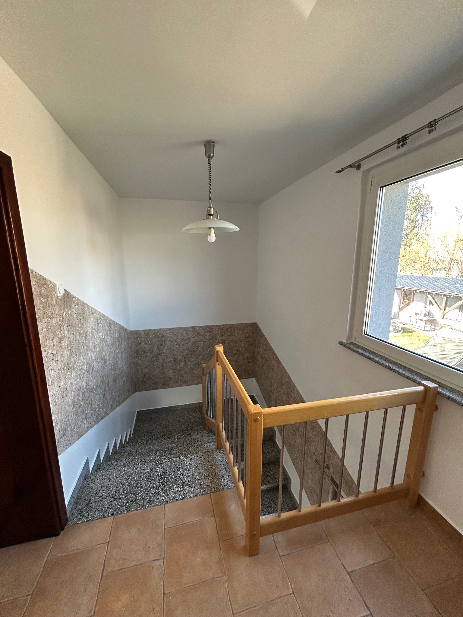 Wnętrze klatki schodowej z terakotową podłogą, schodami wykończonymi lastryko i ścianami z dekoracyjną tapetą korkową do połowy wysokości, oświetlonej wiszącą lampą z białym kloszem.