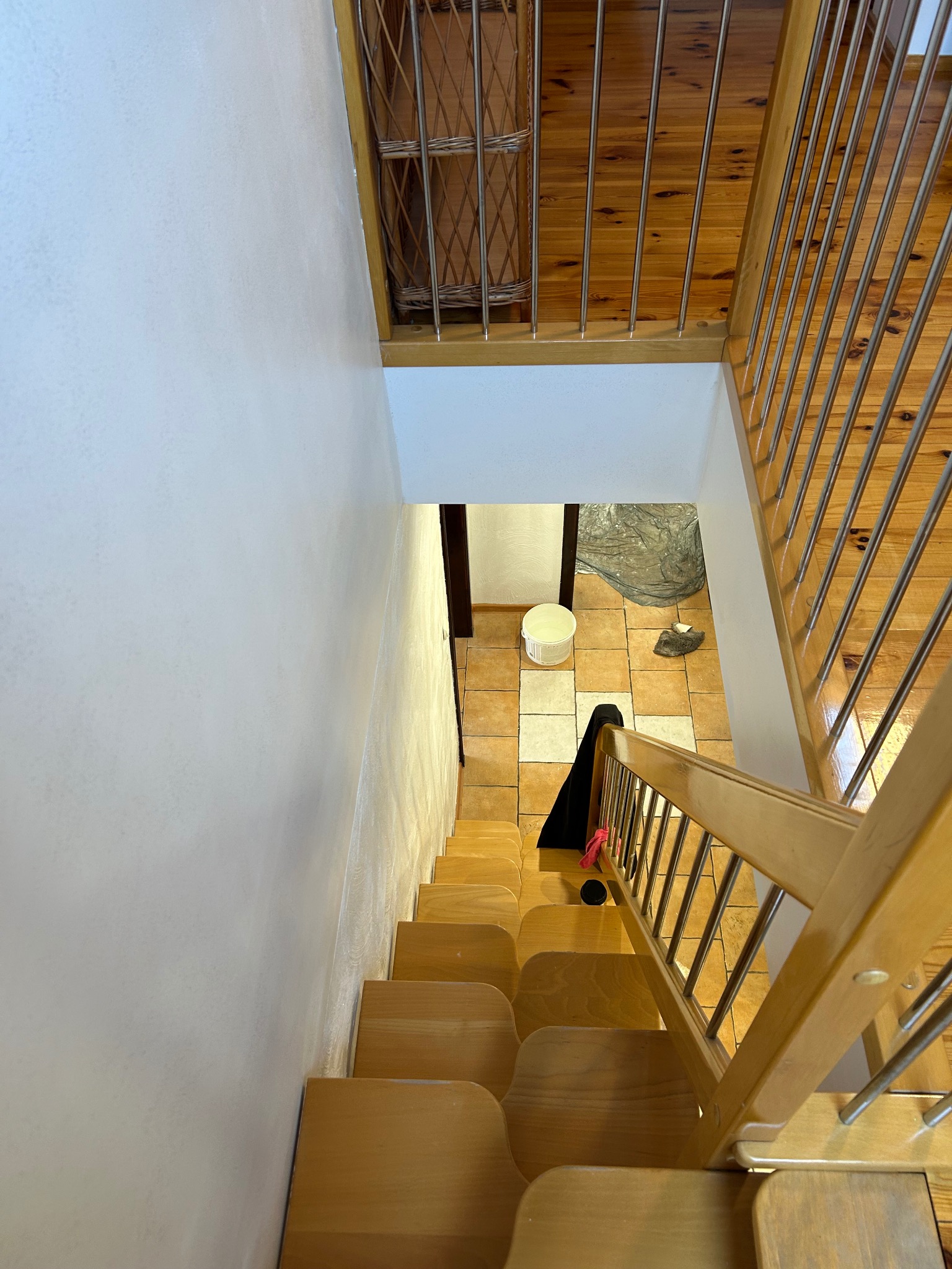 Widok z góry na drewniane schody z metalową balustradą, prowadzące do pomieszczenia z płytkami podłogowymi, wiadrem i folią ochronną, sugerujące prace wykończeniowe.