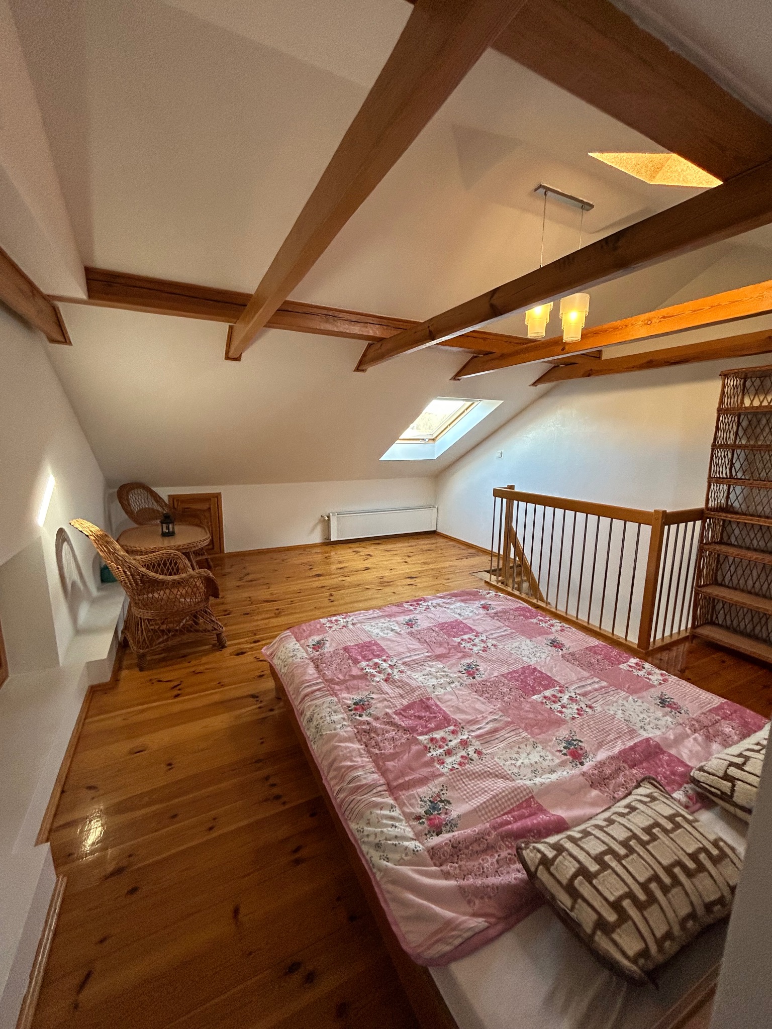 Adaptacja poddasza na sypialnię z drewnianą podłogą, widocznymi belkami stropowymi, oknem dachowym i wiklinowymi meblami.