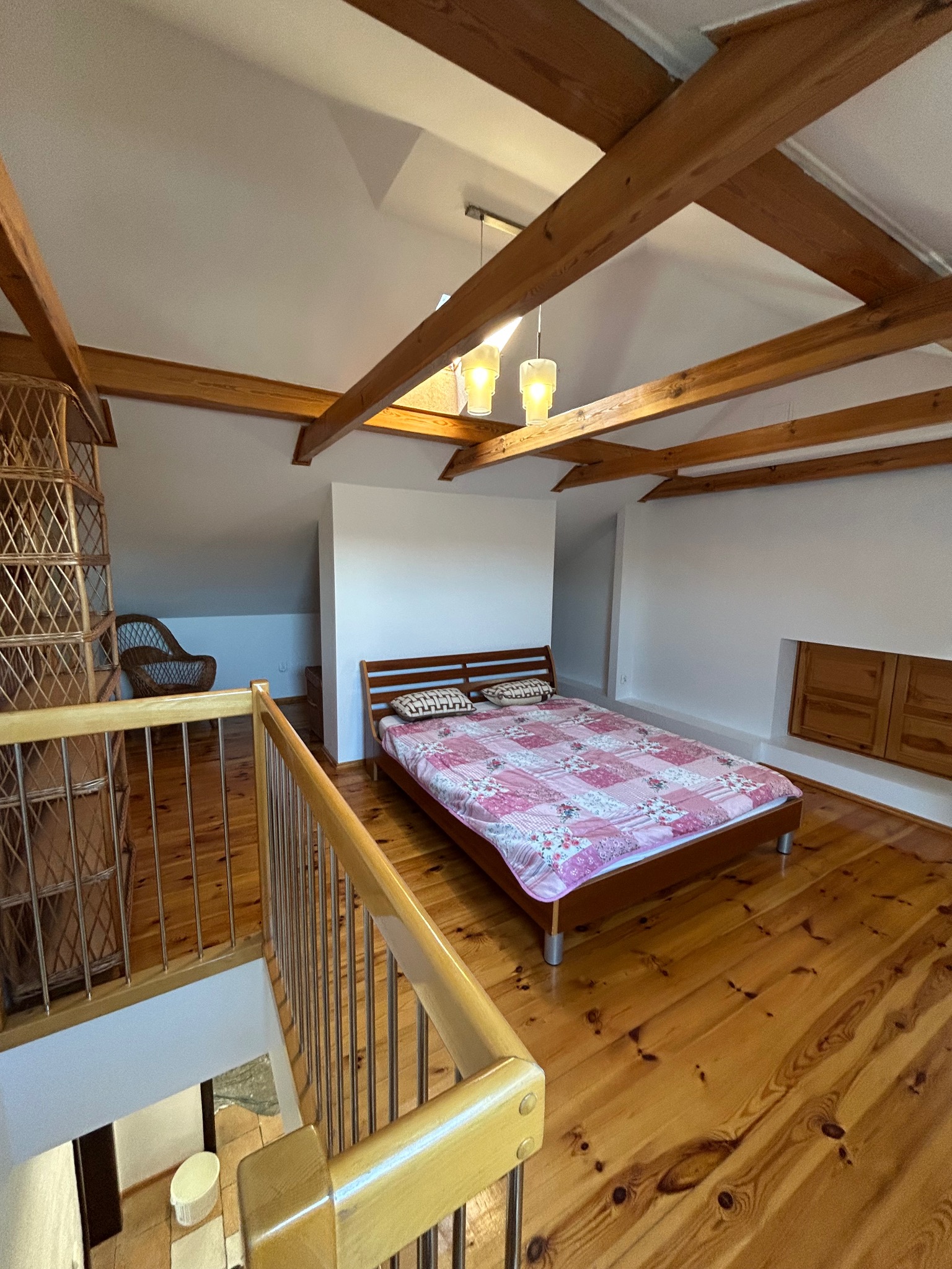 Pokój na poddaszu z drewnianą podłogą, odsłoniętymi belkami stropowymi, łóżkiem z patchworkową narzutą i wiklinowym regałem, widziany z góry z poziomu balustrady schodów.