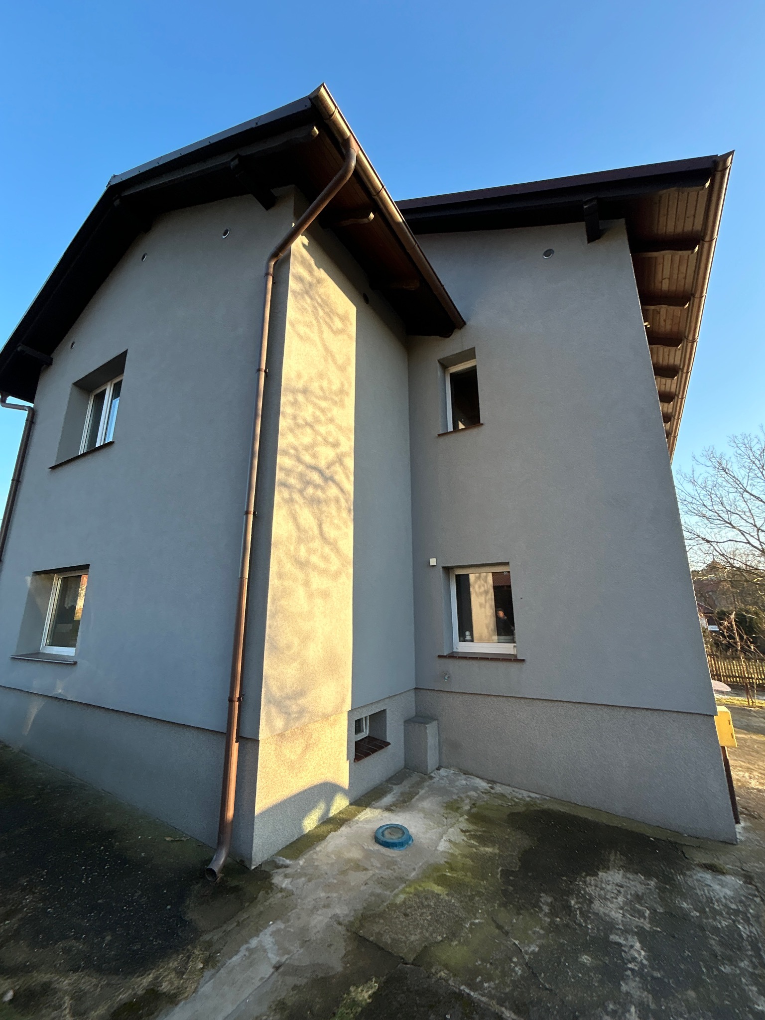 Dwukondygnacyjny dom z szarą elewacją, brązowymi rynnami i drewnianą podbitką dachową, widziany pod kątem w słoneczny dzień.