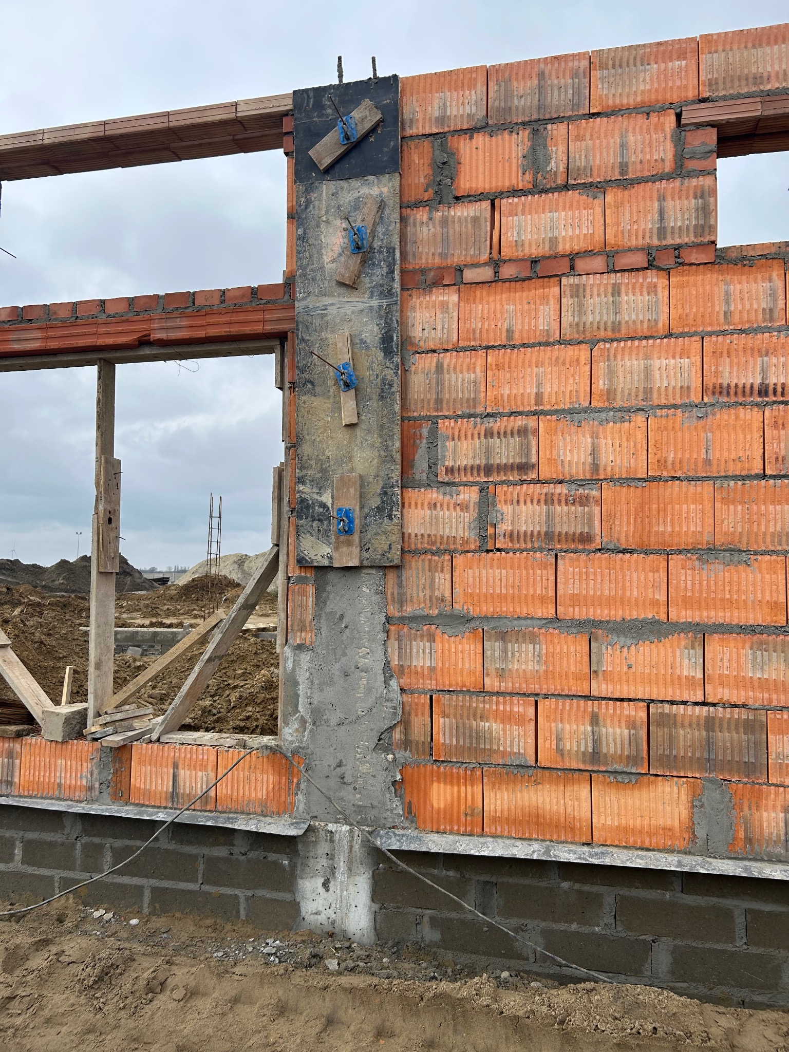 Wznoszenie ściany z czerwonej cegły ceramicznej z widocznymi otworami okiennymi, z użyciem drewnianych elementów szalunkowych i metalowych kotew, na tle placu budowy i pochmurnego nieba.