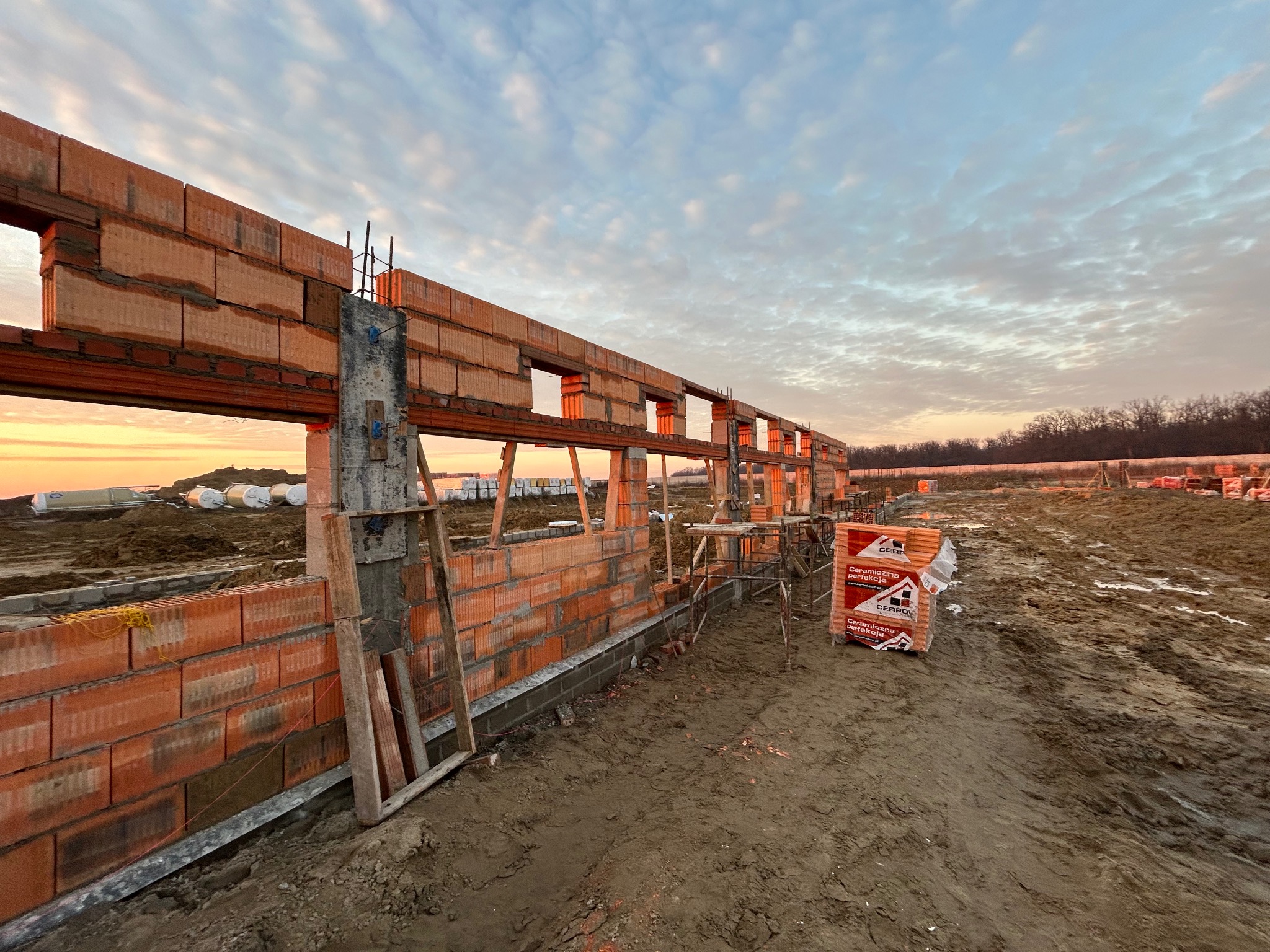Wznoszenie muru z czerwonej ceramiki poryzowanej na fundamencie w trakcie budowy domu, widoczne rusztowania i paleta z bloczkami, tło stanowi zachodzące słońce i częściowo zachmurzone niebo.