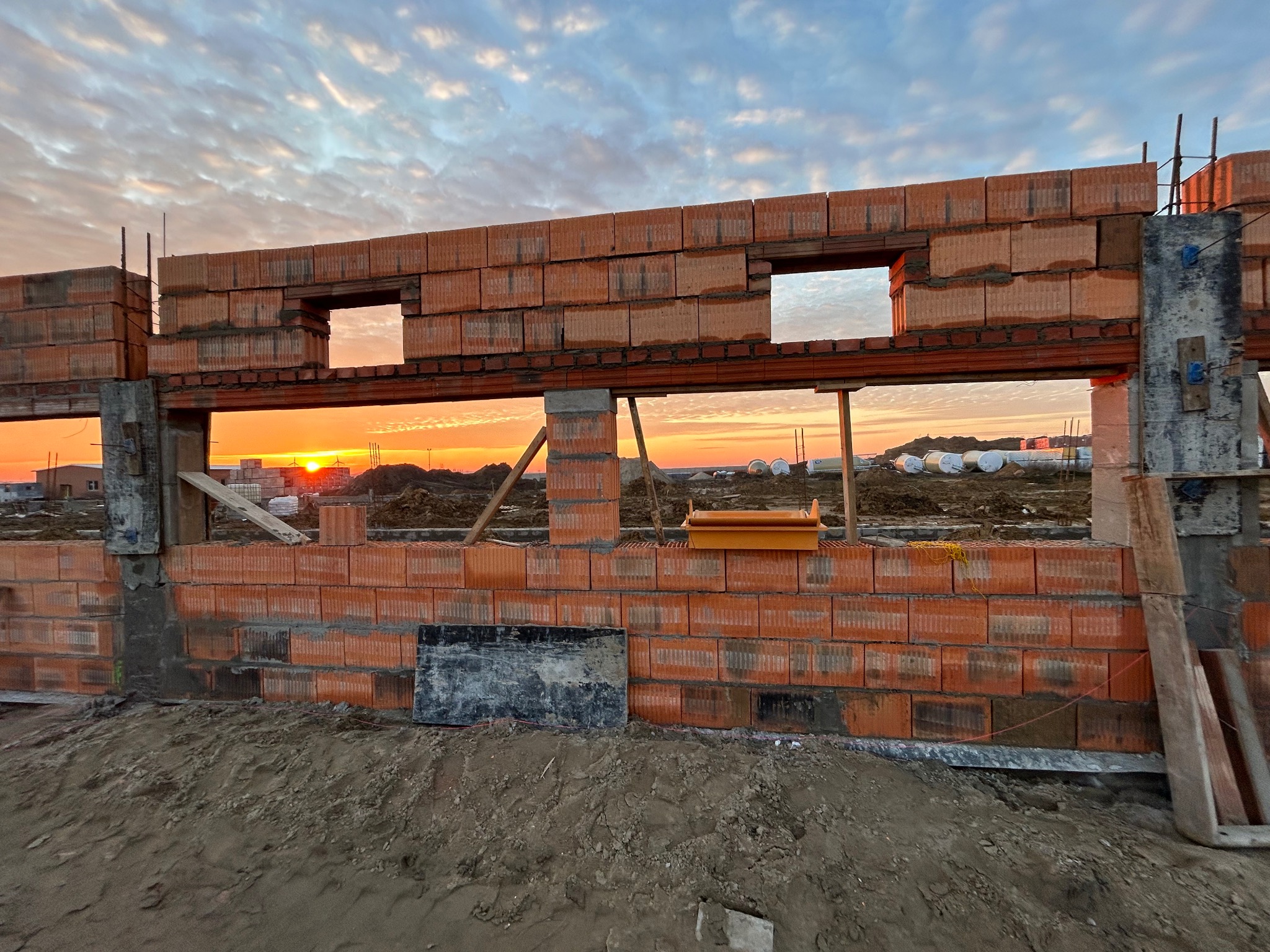 Wznoszone ściany domu murowanego z czerwonej cegły ceramicznej w trakcie budowy, widoczne otwory okienne i słupki konstrukcyjne, na tle zachodzącego słońca i częściowo zachmurzonego nieba.
