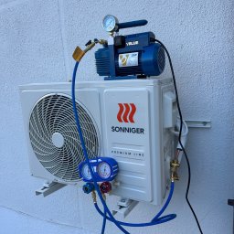 Climate Crafters - Biała jednostka zewnętrzna klimatyzacji Sonniger Premium Line, podłączona do niebieskiej pompy próżniowej VALUE z manometrem, zamontowana na białej, teksturowanej ścianie.