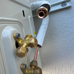 Climate Crafters - Szczegółowe ujęcie złącza klimatyzacji z miedzianymi rurami i izolacją, przymocowanego do białej ściany. Widoczne śruby mocujące i fragment obudowy klimatyzatora.