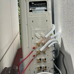 Climate Crafters - Montaż klimatyzacji: tylny panel jednostki zewnętrznej z podłączonymi rurami miedzianymi i przewodami, widok z bliska na instalację w przestrzeni między budynkami.