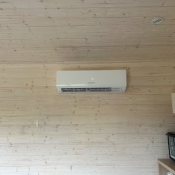 Climate Crafters - Biała klimatyzacja Sonniger na drewnianej ścianie w jasnym pomieszczeniu z lampą i piekarnikiem na szafce. Widoczne oświetlenie sufitowe.