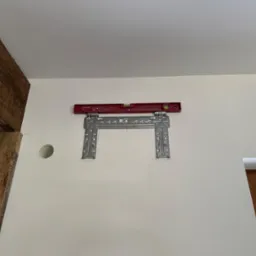 Montaż wspornika pod klimatyzator z poziomicą na ścianie w Gdańsku. Widoczny otwór na instalację. Fragment drewnianej belki stropowej. Okno z zielenią w tle.